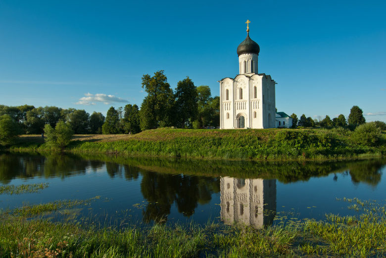Храм Покрова на Нерли на фоне голубого неба, отражающийся в воде среди зелени лугов.