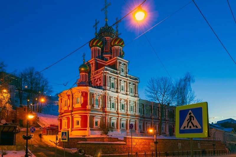Строгановская церковь в Нижнем Новгороде в вечернее время зимой, с подсветкой фасада и заснеженной улицей на переднем плане.